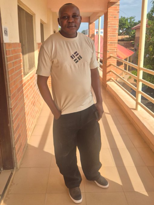 A man stands on an outdoor tiled walkway or balcony, wearing a beige 'CHRISTIAN' t-shirt with a geometric logo, dark pants, and gray shoes, with brick pillars and a view of other buildings in the background.