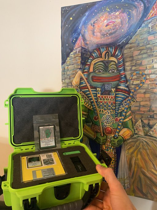A person holds a bright green protective case containing what appears to be an Amstrad CPC computer cartridge, displayed in front of an Egyptian-themed painting.