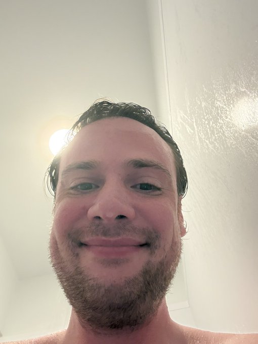 A close-up selfie of a smiling man with wet dark hair and a beard, appearing to be in a shower with white walls and a light visible in the background.