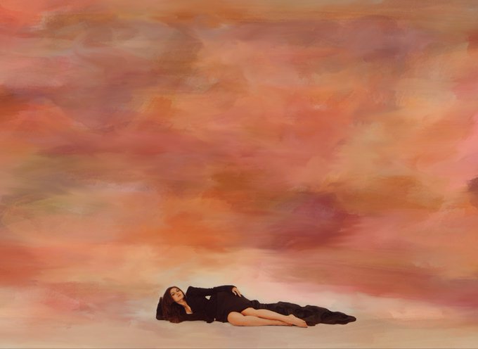 A woman in a black dress lying down against a painted backdrop of swirling pink, orange, and beige hues resembling a cloudy sunset.