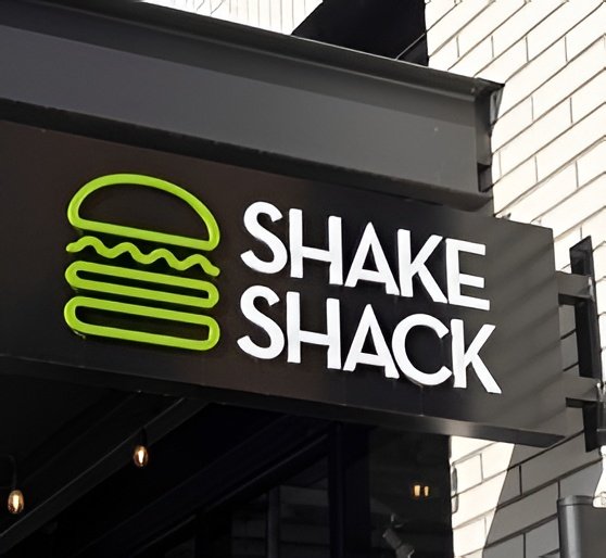 A storefront sign for the restaurant chain 'SHAKE SHACK' displays their green burger logo next to white lettering on a black background.