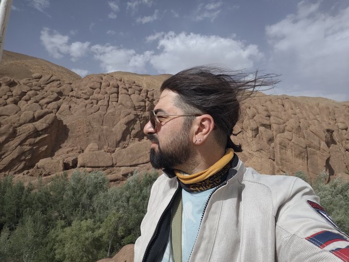 The image features a man with long hair wearing sunglasses, a white jacket, and a scarf around his neck. He appears to be standing near some mountains or rock formations, possibly at a scenic location like Morocco. The photo has been captioned "Gm from Morocco", indicating that it was taken in the country of Morocco by someone who is also present in the image.

The man's attire and surroundings suggest he might be on an outdoor adventure or exploring nature. His sunglasses could indicate that the weather is sunny, which adds to the overall atmosphere of a pleasant day spent outdoors. The presence of mountains or rock formations further emphasizes the natural setting where this scene takes place.