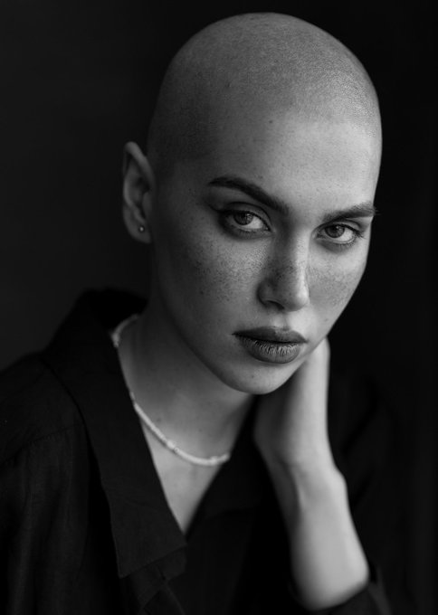 A black and white portrait of a bald woman wearing a dark collared shirt and a delicate necklace, gazing intensely at the camera with freckled skin and dramatic makeup.
