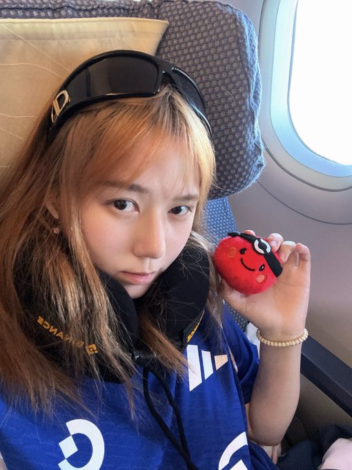 A young woman takes a selfie on an airplane, wearing sunglasses on her head and a blue shirt with 'GAINWARD' text, while holding up a small red plush toy.