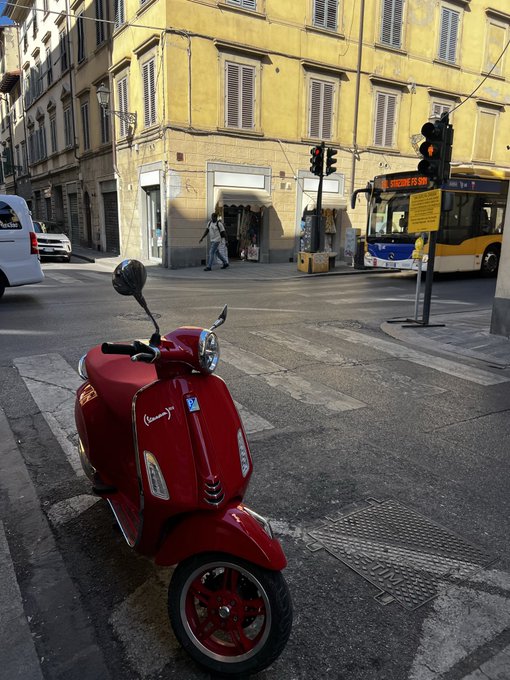 A street scene showing a red Vespa-style scooter parked at a crosswalk in what appears to be an Italian city, with a yellow building, traffic lights, and an orange and white bus in the background.