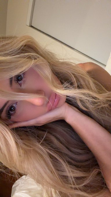 A close-up selfie of a young woman with long blonde hair, resting her face on her hands with heavy eye makeup, taken in soft lighting.