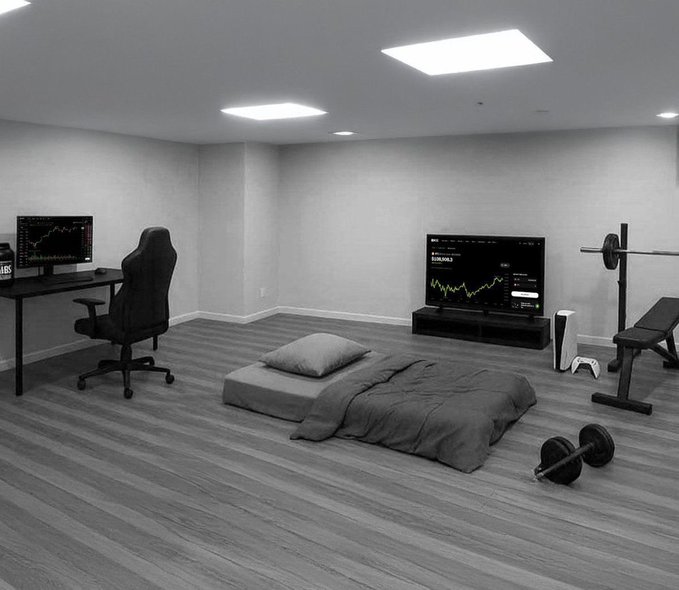 A minimalist gray room featuring a gaming setup with a desk and monitor on the left, a TV displaying financial charts in the center, a floor mattress, and weightlifting equipment on the right.