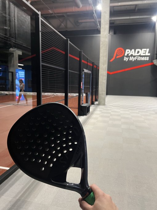 A hand holds a black padel racket in front of an indoor padel court with a 'PADEL by MyFitness' logo on the wall.