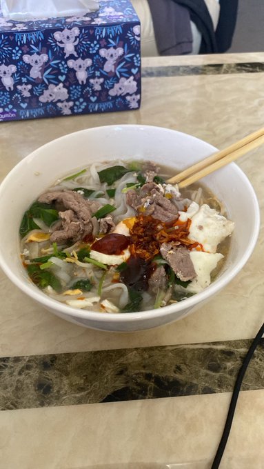 A bowl of Vietnamese noodle soup, likely pho, topped with beef, egg, chili oil, and greens sits on a marble counter with chopsticks resting on the rim.