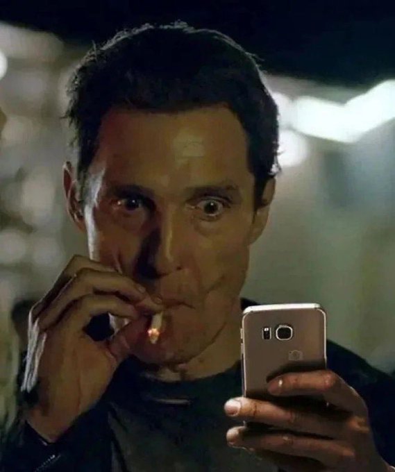 A man lighting a cigarette while intensely looking at a smartphone, with a shocked expression on his face.
