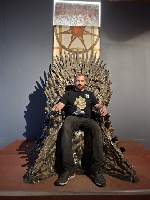 A bearded man wearing a black t-shirt with a cartoon graphic sits on a throne made of swords, resembling the Iron Throne from Game of Thrones, displayed behind glass.