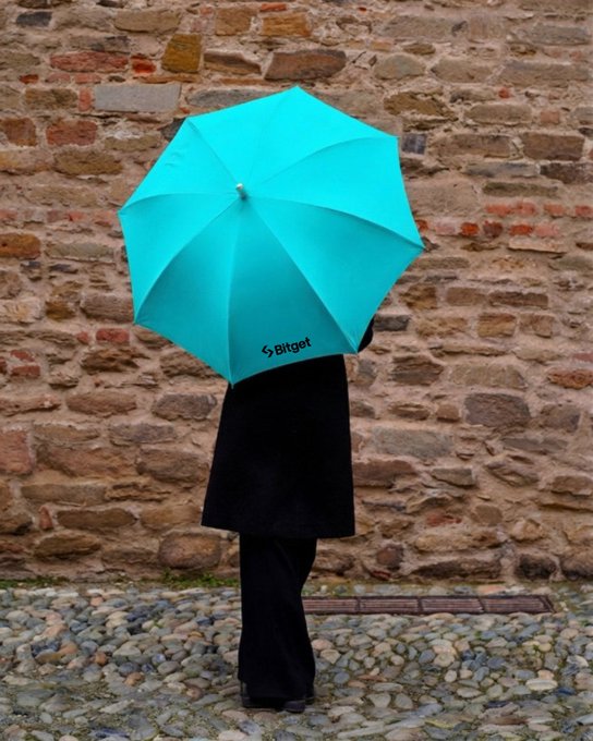 A person in a black coat stands with their back to the camera holding a bright turquoise umbrella branded with 'bitget', against an old stone wall.