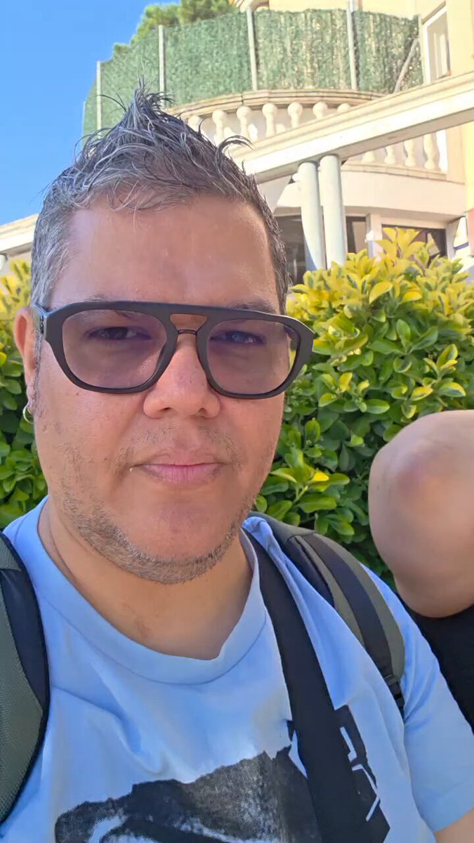 The image features a man wearing glasses, sunglasses, and a t-shirt with a picture of a person's face on it. The man has short hair and is smiling at the camera. In the background, there are some buildings visible behind him. The caption of the image reads "¡Saliendo! Hoy #Zcast y la comunidad de #Zcash en Español estará presente en el @bcc8333 #Zcash". This caption is related to a post on social media, possibly about an event or gathering in Barcelona. The man's attire and the presence of buildings suggest that he might be attending such an event or exploring the city.