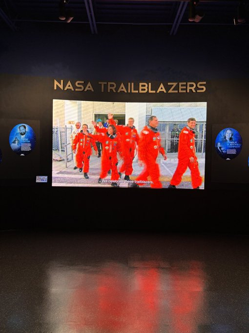 An exhibit at a NASA museum or facility titled 'NASA TRAILBLAZERS' displaying a video of astronauts in orange flight suits on a dark wall with smaller circular portraits on either side.