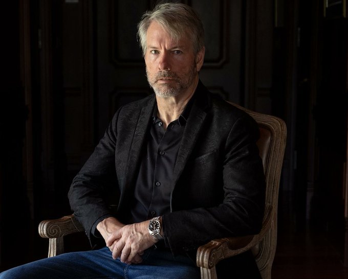 Michael Saylor, executive chairman of Strategy (formerly MicroStrategy), sits in a wooden armchair wearing a black blazer and dark shirt, with a watch visible on his wrist. The image likely accompanies a post about his recent ambiguous Sunday chart commentary following Strategy's largest Bitcoin sale of 3,588 BTC for $216 million.