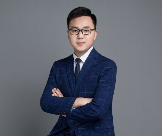 A professional headshot of an East Asian man wearing glasses, a navy blue windowpane-check suit, and a dark tie, posing with his arms crossed against a gray background.