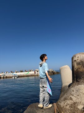 The image features a young man standing near water, possibly at a pier or dock. He has his arms outstretched towards the sky, giving off an impression of wonder and awe as he looks up into the vast expanse above him. The photo is taken from a low angle, which emphasizes the height of the person's arms and adds to the sense of grandeur in the scene.