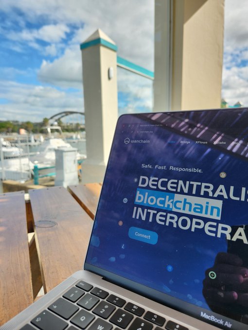 A MacBook Air laptop sits on a wooden outdoor table, displaying the Wanchain website with text including 'Safe. Fast. Responsible.', 'DECENTRALIZED', 'blockchain', and 'INTEROPERA' along with a 'Connect' button, with a marina visible in the background.