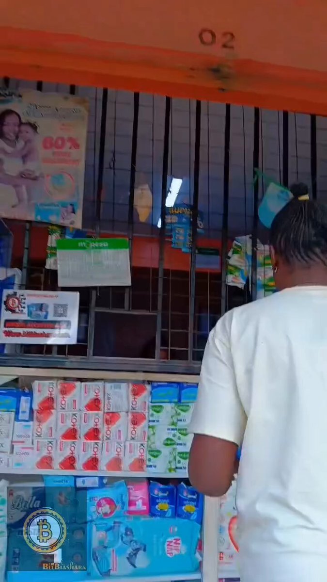 The image shows a person standing next to a store display filled with various products, including diapers and other items for sale. The individual appears to be browsing through the selection or possibly making a purchase. The store has an orange door that serves as its entrance, and there is a sign on it advertising diapers. A post on social media also mentions the presence of diapers in the store's display window.