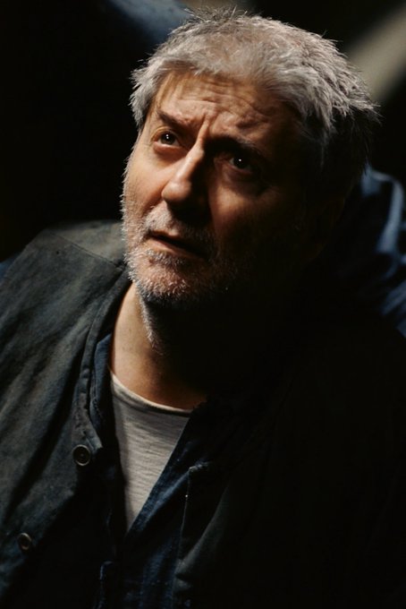 The image features a man with gray hair wearing a blue jacket, looking down at his hands or feet while sitting in front of him. The background appears to be dark, which emphasizes the subject's presence. There are no texts present in the image, so we can only see what is directly related to the main subject and not any additional information about the post.