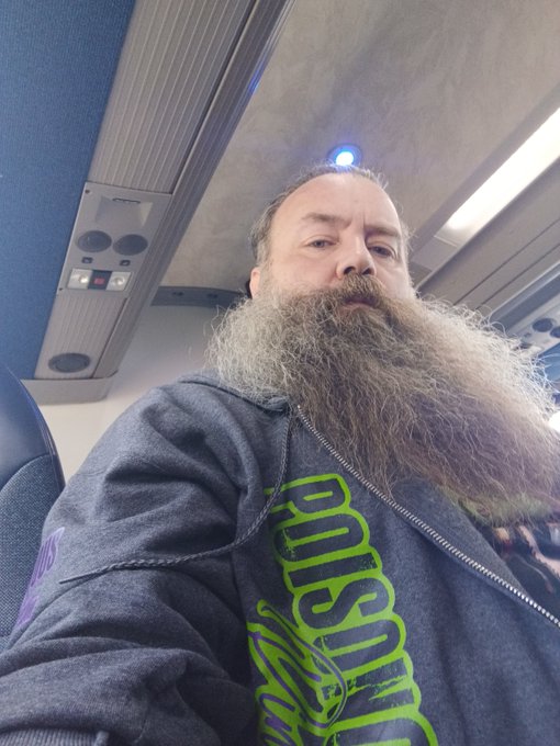 A bearded man in a dark gray hoodie with green lettering sits on what appears to be an airplane or train, looking directly at the camera.