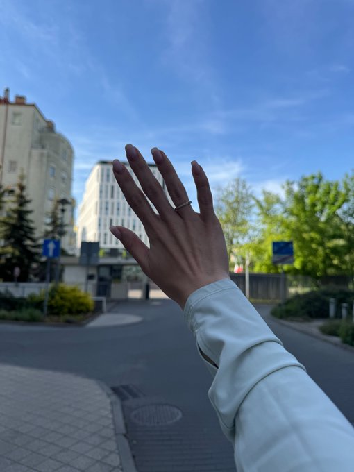 A person's hand raised against a backdrop of a blue sky, trees, and buildings, with a ring visible on the ring finger.