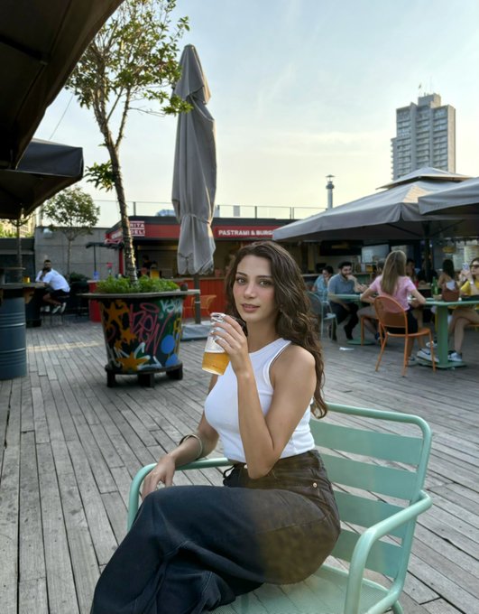 A woman with long dark hair sits at an outdoor cafe holding a glass of beer, with a tall building visible in the background.