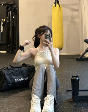 A gym selfie of a woman in athletic wear flexing her arm while sitting on the floor, with a yellow punching bag and a 'GGate VIP' duffel bag nearby.