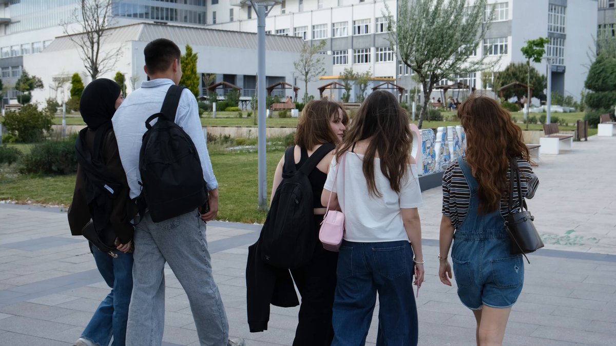 The image shows a group of five people walking down a sidewalk, with four of them carrying backpacks while one person has a handbag. They are all dressed similarly, suggesting they might be students or part of an organized event. The background features a building, indicating that they are in an urban setting. The post about the university tour seems to be promoting blockchain education and encouraging people to support it by sharing the image on social media platforms.