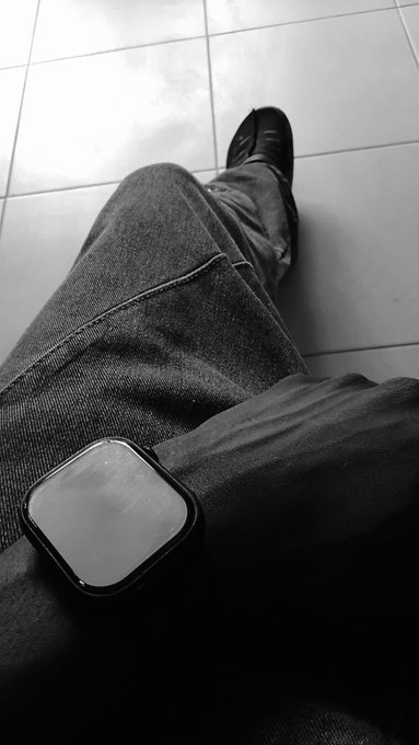 A black-and-white photo from a first-person perspective looking down at a person's legs in jeans and dark shoes, with a smartwatch resting on their lap.