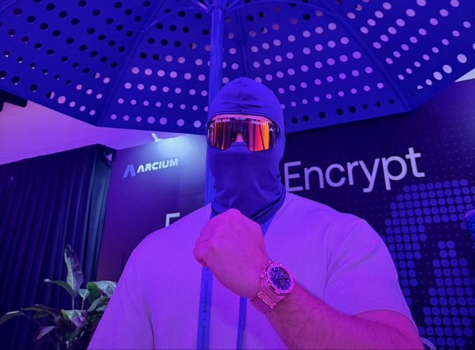 The image features a man wearing a mask, standing under an umbrella with his hand on his face. The background of the photo shows a banner that says "Encode" written in white letters. The man appears to be posing for a picture or attending an event related to the Encode platform.