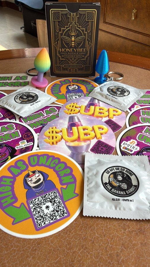 Adult novelty products displayed on a table, including a 'HONEY BEE ROYALE' vibrating ring box, rainbow and blue butt plug items, and branded merchandise stickers and coasters with QR codes featuring slogans like 'GREASE AS UNICORN' and 'RIDE OR GO HOME - THE BANANA BANDIT'.