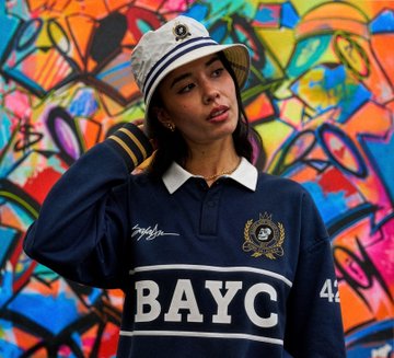 A woman wearing a BAYC polo shirt and a white captain's hat poses against a colorful graffiti wall, promoting the upcoming BAYC x King Saladeen Anchor Drop and burgee flag raffle scheduled for June 30th, 2026.
