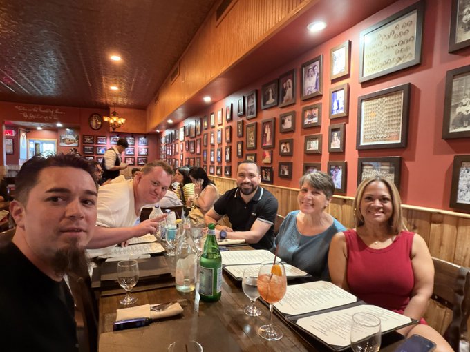 A group of five people sit together at a wooden table in a restaurant with red walls covered in framed pictures, posing for a photo with drinks in front of them.