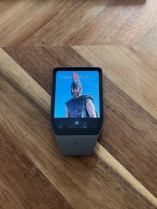 A black smartwatch rests on a wooden floor, displaying the Spotify screen for "Trezor Safe 7" with album artwork featuring a hooded figure, complementing the poster's enthusiastic "yeah loved it" comment about the music.