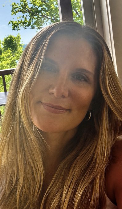 A close-up selfie of a woman with long brown hair, smiling indoors with a window and greenery visible in the background.