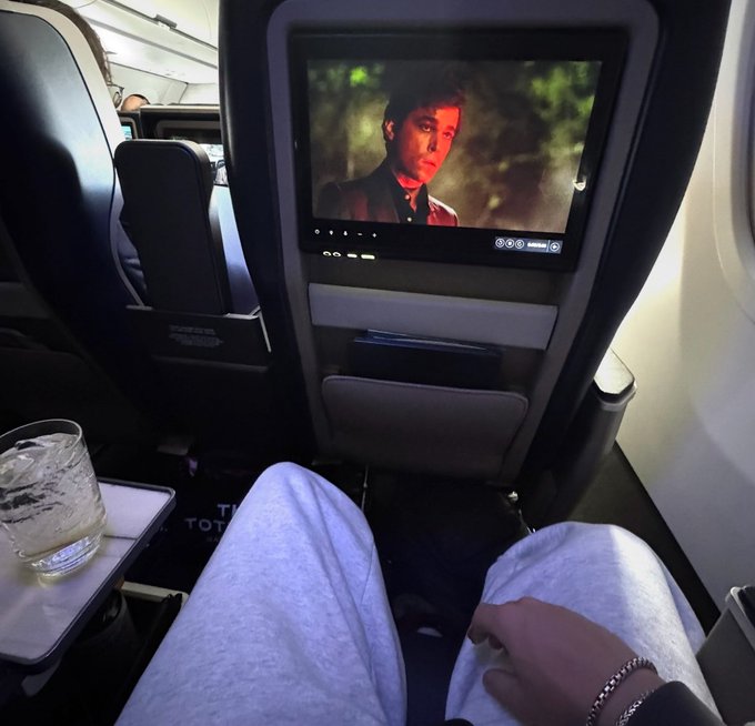 A first-person view from an airplane seat showing the in-flight entertainment screen playing a movie, with a glass of water on the tray table.