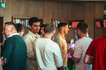 A group of people socializing at what appears to be an indoor event or gathering, with bookshelves visible in the background.