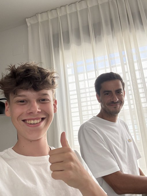 Two smiling individuals pose for a selfie in a bright room with sheer white curtains, both giving enthusiastic thumbs up while wearing white shirts—one with partially visible gold lettering. The post's "CARLOS X LEON SEASON" caption suggests this captures the two named subjects back together for a new collaboration or content series.