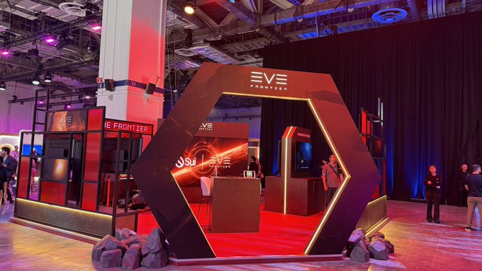 An exhibition booth for "EVE FRONTIER" featuring a futuristic hexagonal entrance with red lighting, product displays, and attendees.