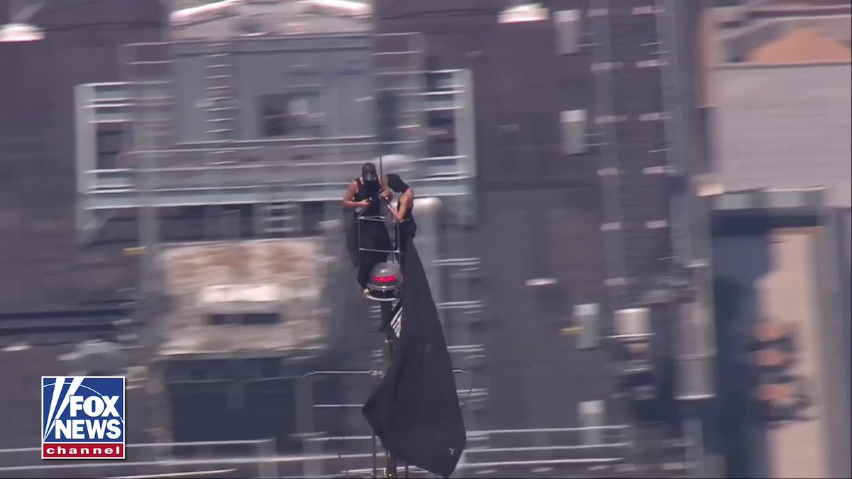 Two people are perched atop a towering structure, unfurling a large black flag against a hazy urban skyline, with a "FOX NEWS channel" logo in the bottom left corner. The image captures a daring, lofty display that visually echoes the post's message about reaching high places to make bold statements.