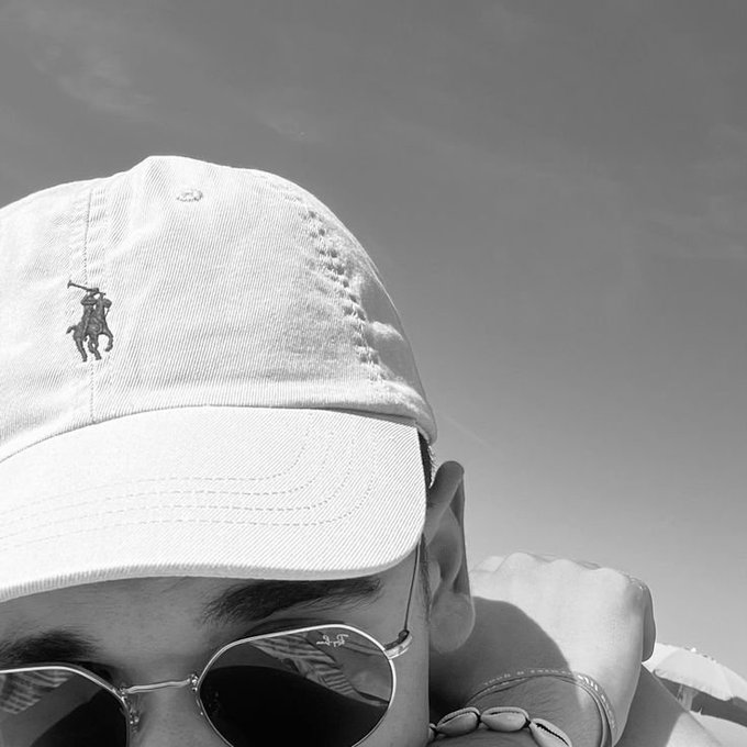 A black and white close-up photo of a person wearing a baseball cap featuring a polo player logo, along with round sunglasses, with another person visible in the background.