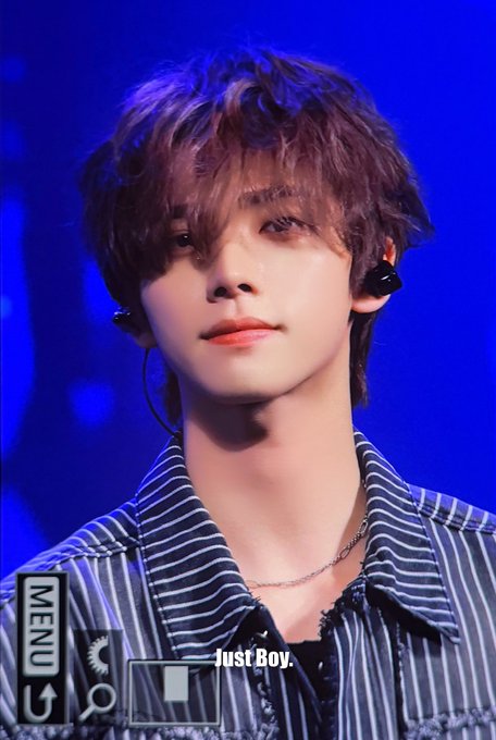 Close-up photo of a young male performer with wavy brown hair wearing a striped denim shirt and in-ear monitors against a blue stage background, watermarked 'JustBoy' with a MENU graphic in the corner.