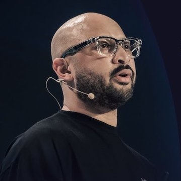 A bald man with a beard and glasses, wearing a black shirt and headset microphone, appears to be giving a presentation or speech.