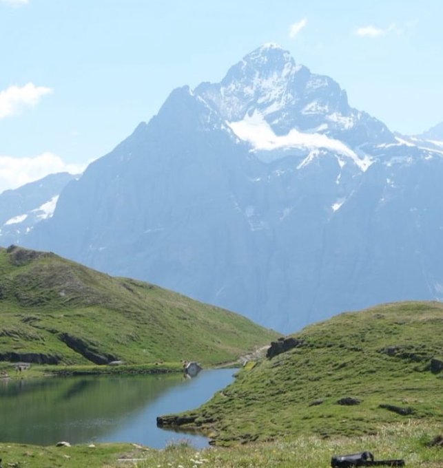 A scenic alpine landscape featuring a snow-capped mountain peak towering over rolling green hills and a small reflective lake under a blue sky.
