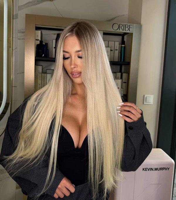 A woman with long blonde hair wearing a black top in a salon, with Oribe and Kevin Murphy product displays visible in the background.