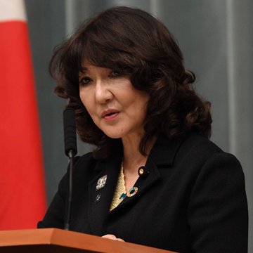 A woman with dark wavy hair, wearing a black blazer with a brooch and pearl necklace, speaks at a podium with a microphone, with a Japanese flag visible in the background. The image appears to show Japan's Finance Minister Satsuki Katayama, who is quoted discussing Japan's plans to legalize cryptocurrency ETFs.
