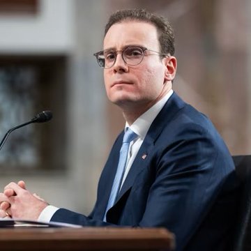 A photograph of a man wearing glasses and a dark suit with a blue tie, sitting in front of a microphone at what appears to be a formal hearing or testimony.