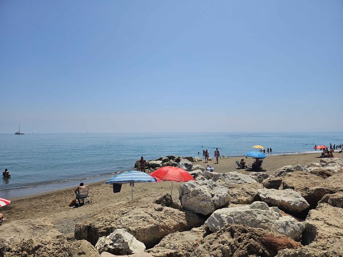 A photograph of a sunny Mediterranean-style beach with clear blue water, people swimming and relaxing under red and blue umbrellas, large white rocks in the foreground, and a sailboat visible on the horizon.