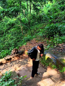 A person wearing dark clothing stands in a lush green forest beside a rocky stream, looking down at the ground.